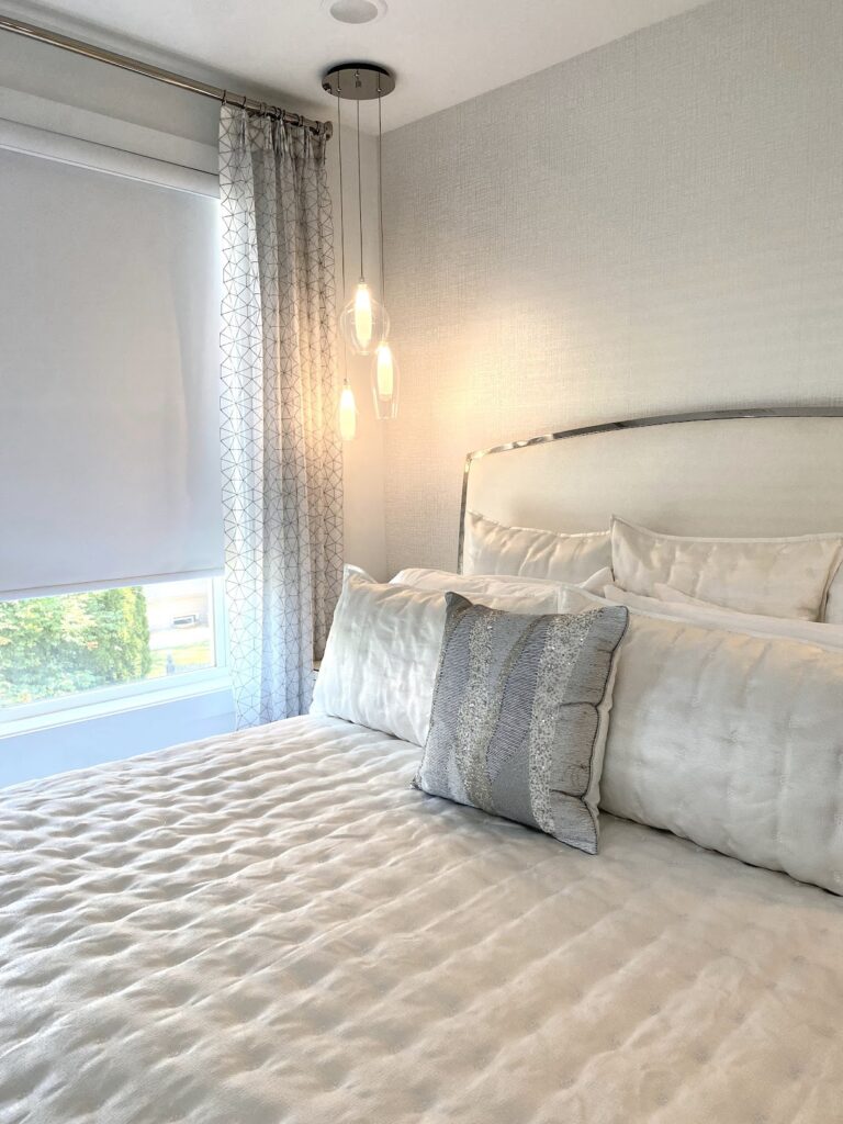 Contemporary master bedroom designed with Bernhardt furniture, luxurious Kravet fabric layered bedding, chrome finish accents, cascading glass pendant lights, and a sleek roller shade — where every detail is intentional