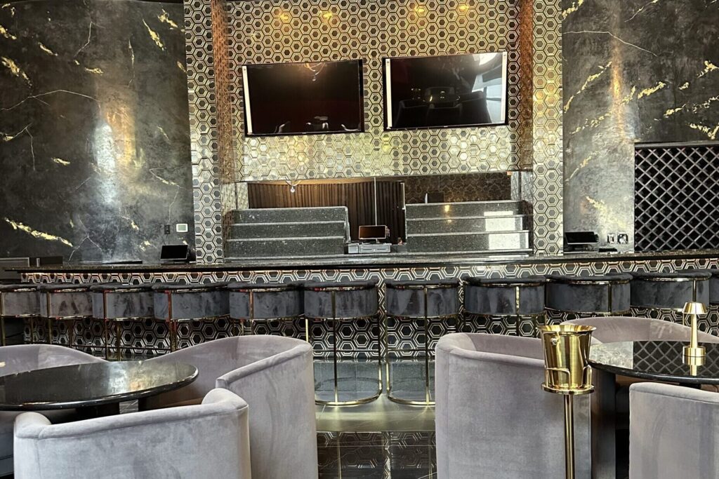 A full view of a custom restaurant and lounge bar with dark marble column accents, a hexagon mosaic tile bar front, gray velvet barrel lounge chairs, dark marble floors, gold accent pieces, and dual wall-mounted TVs — luxury hospitality design done right