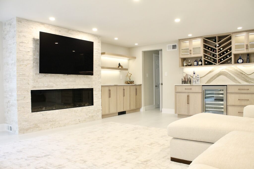 An open concept luxury basement renovation showcasing a stacked stone fireplace wall with wall-mounted TV, warm under-cabinet lighting, light oak built-in cabinets, and a custom dry bar with Cambria Brittanicca Warm backsplash and chevron wine rack — the ultimate entertainment space