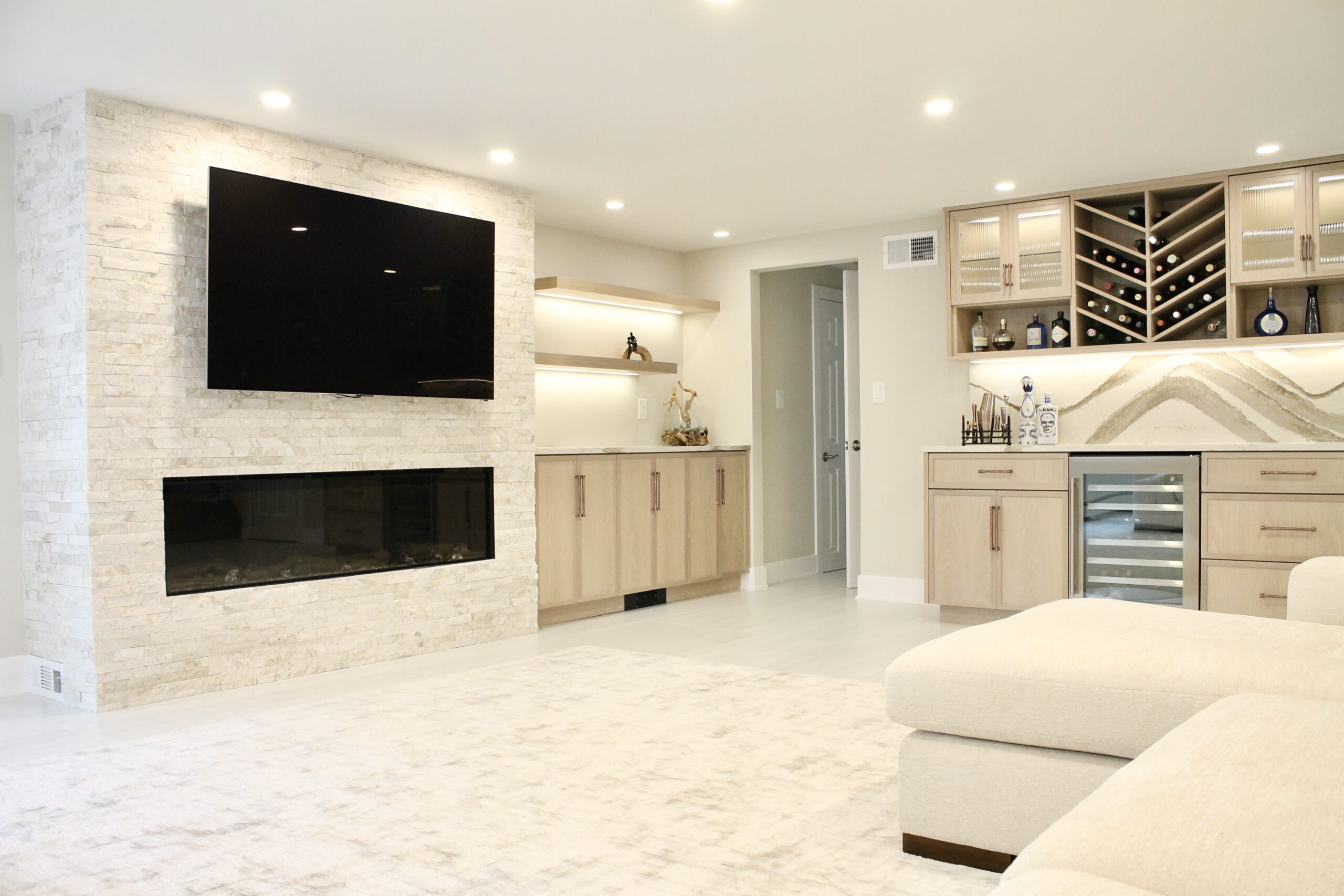 An open concept luxury basement renovation showcasing a stacked stone fireplace wall with wall-mounted TV, warm under-cabinet lighting, light oak built-in cabinets, and a custom dry bar with Cambria Brittanicca Warm backsplash and chevron wine rack — the ultimate entertainment space