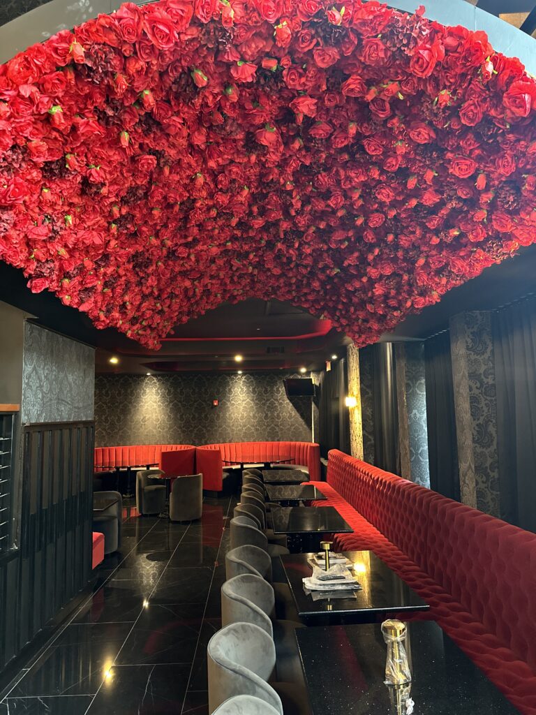 A jaw-dropping luxury restaurant dining room featuring a massive red rose ceiling installation cascading over red tufted velvet banquette seating, gray velvet barrel chairs, black marble floors, damask wallpaper walls, and dramatic moody lighting — a room that stops you in your tracks