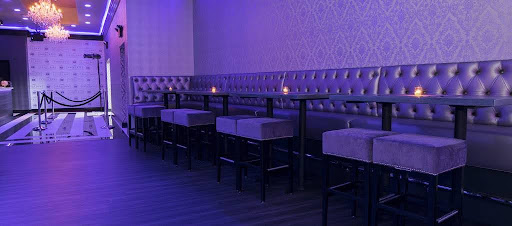 Moody venue with floor-to-ceiling tufted silver banquette seating, high-top tables with upholstered stools, dramatic purple ambient lighting, and a red carpet step-and-repeat backdrop — equal parts VIP lounge and event-ready glam