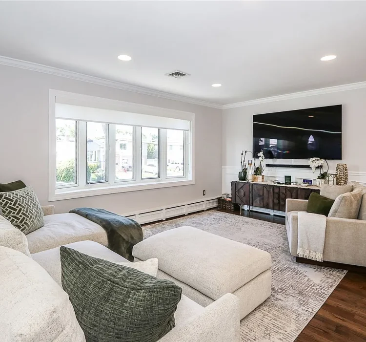 Modern living room design featuring a plush neutral sectional sofa with green and white throw pillows, large wall-mounted TV, geometric dark wood media console, wainscoting accent wall, and hardwood floors